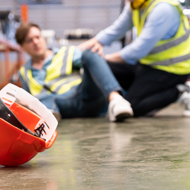 Ouvrier au sol après un accident de travail, assisté par un collègue. Casque de sécurité orange au premier plan, sol industriel.