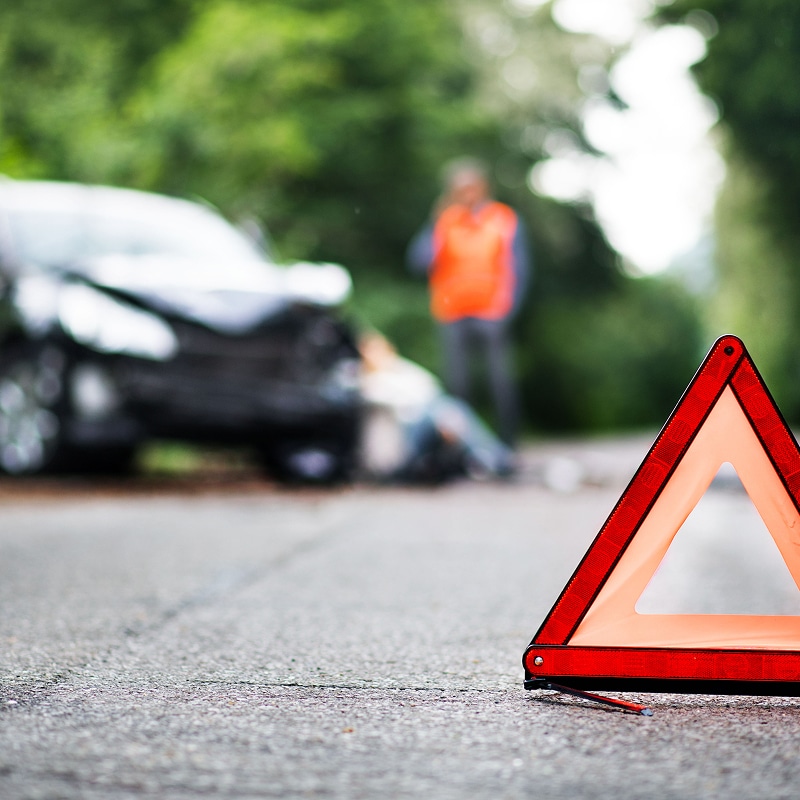 Triangle de signalisation rouge sur route. Accident de voiture avec véhicule noir endommagé, personne au sol, et homme en gilet orange appelant à l'aide.