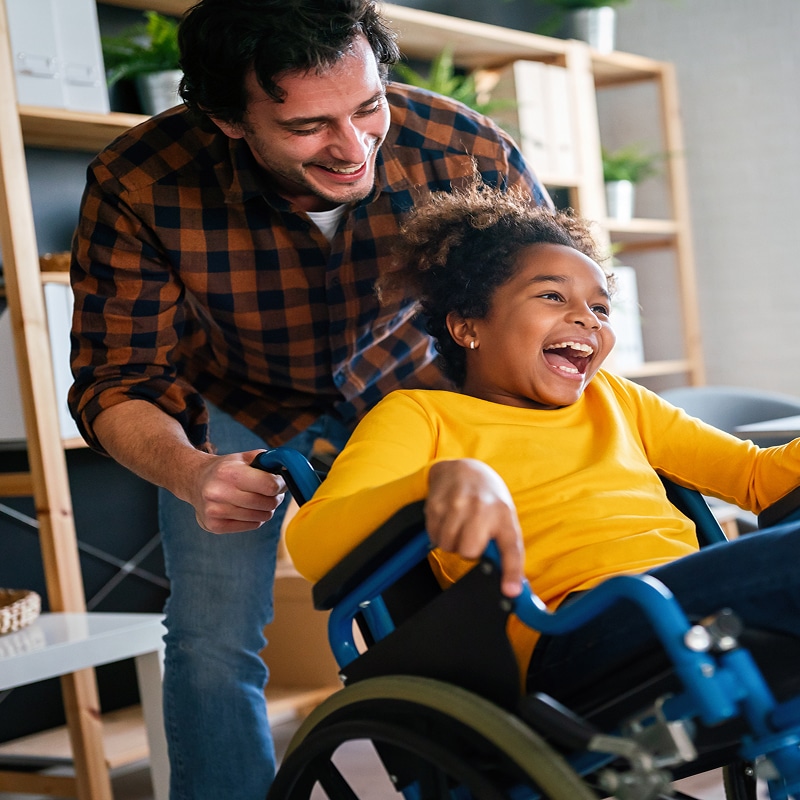 Homme souriant poussant une petite fille riante en fauteuil roulant, partageant un moment de joie.