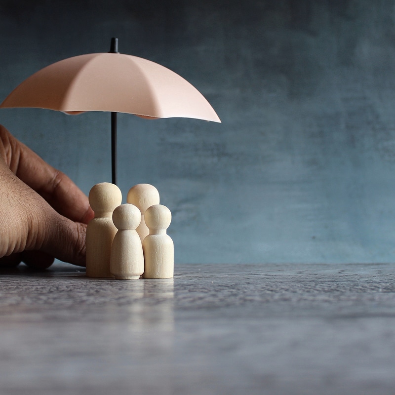 Main protégeant une famille de figurines en bois sous un parapluie rose. Concept de protection familiale ou assurance.