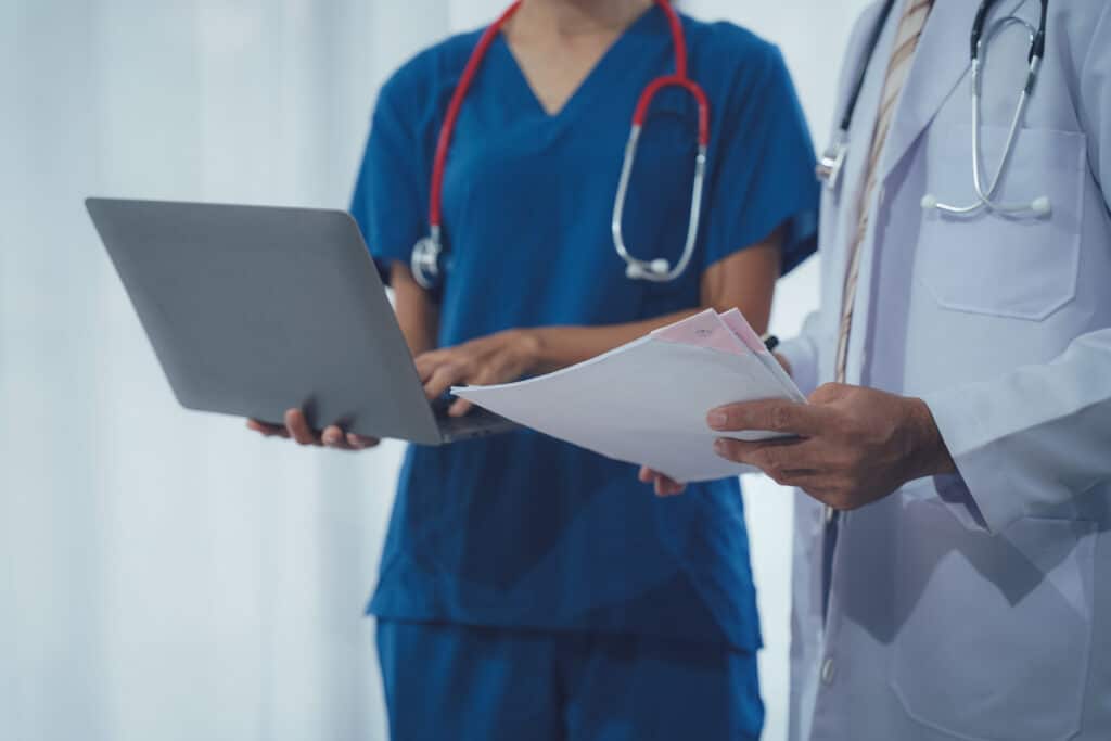 Deux professionnels de santé. Infirmière en scrubs bleus avec stéthoscope utilise un ordinateur portable, un médecin en blouse lit des documents.