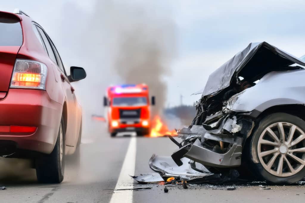 Accident de voiture grave avec épave frontale en feu sur route, pompier et véhicule rouge en arrière-plan. Urgence routière.