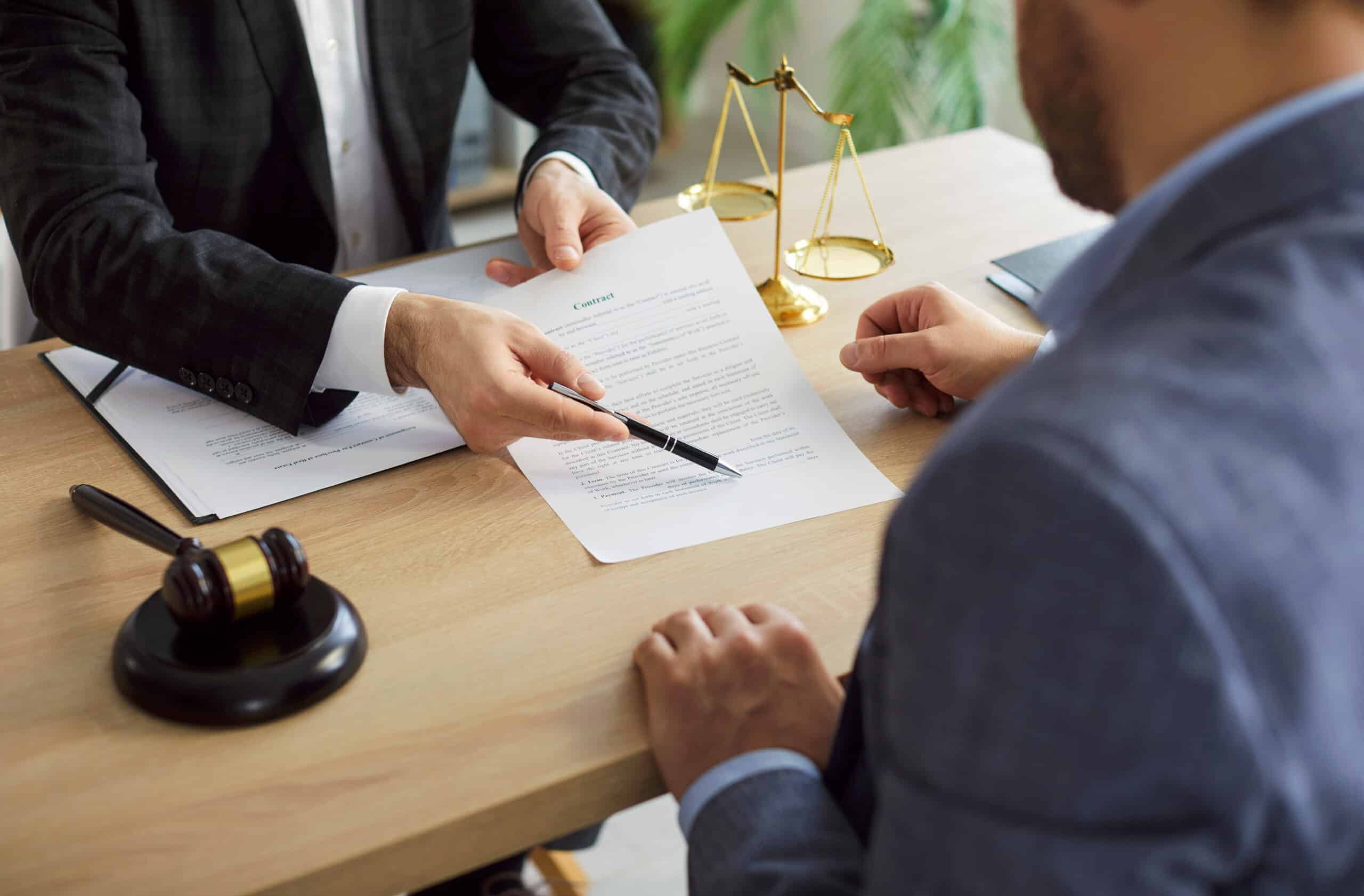 Avocat montrant un contrat à un client avec un stylo sur un bureau en bois. Balance de justice et marteau de juge sont visibles.