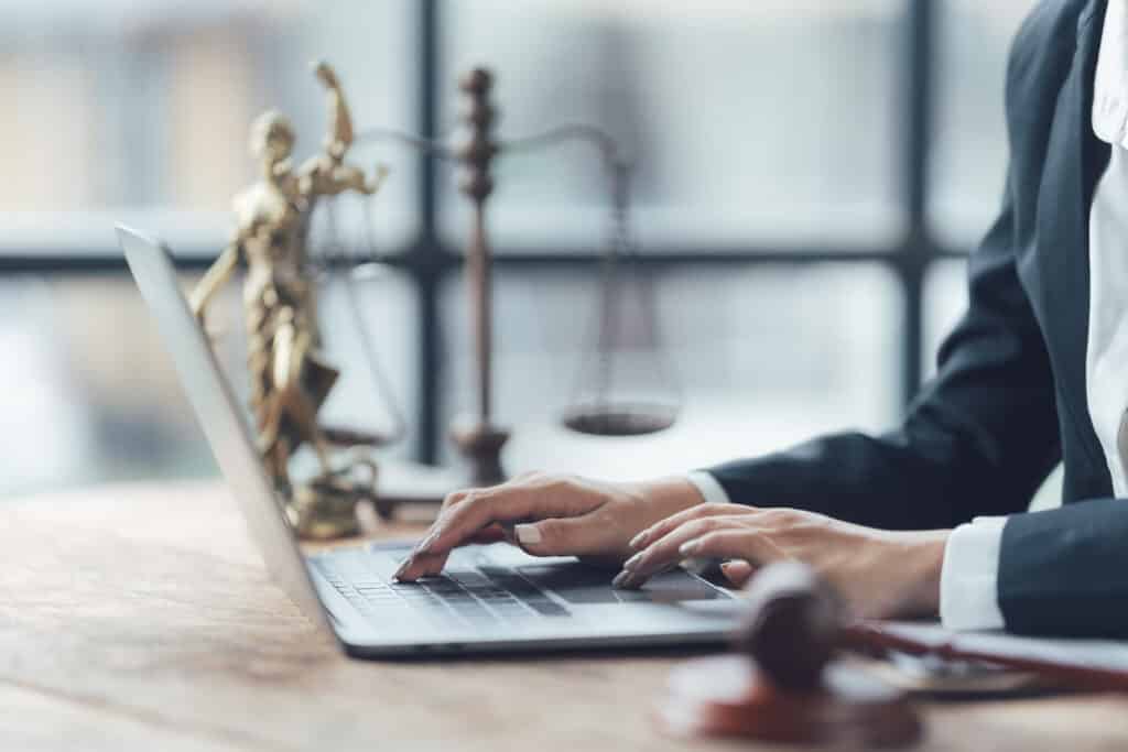 Mains d'un avocat tapant sur un ordinateur portable sur un bureau en bois. Statue de la Justice et marteau de juge en arrière-plan.