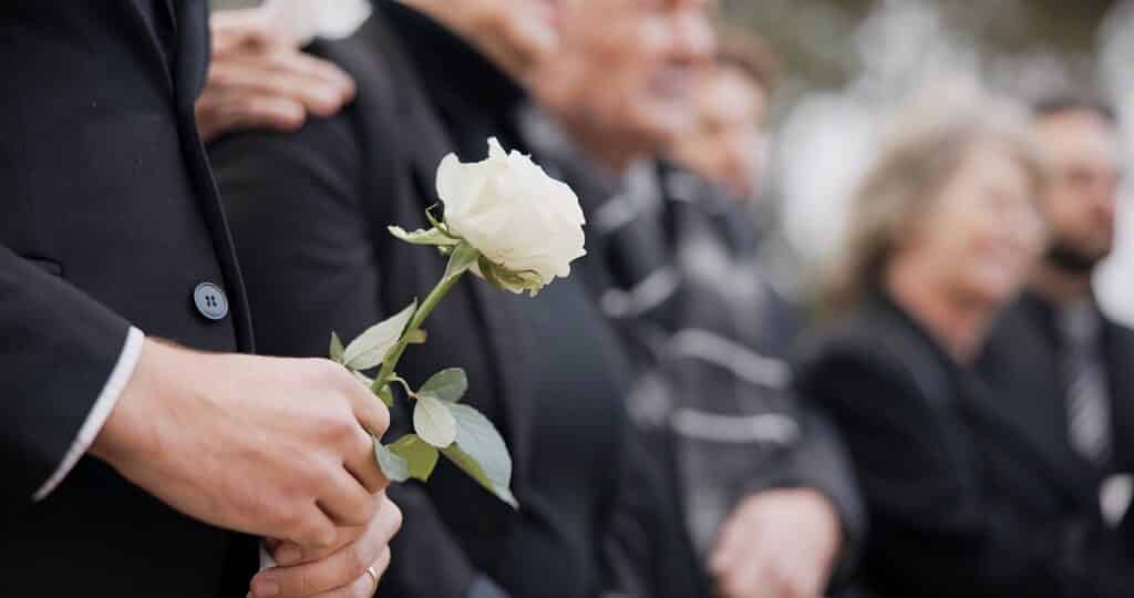 Homme en costume noir tenant une rose blanche, main avec alliance. Public flou, ambiance solennelle lors d'obsèques.