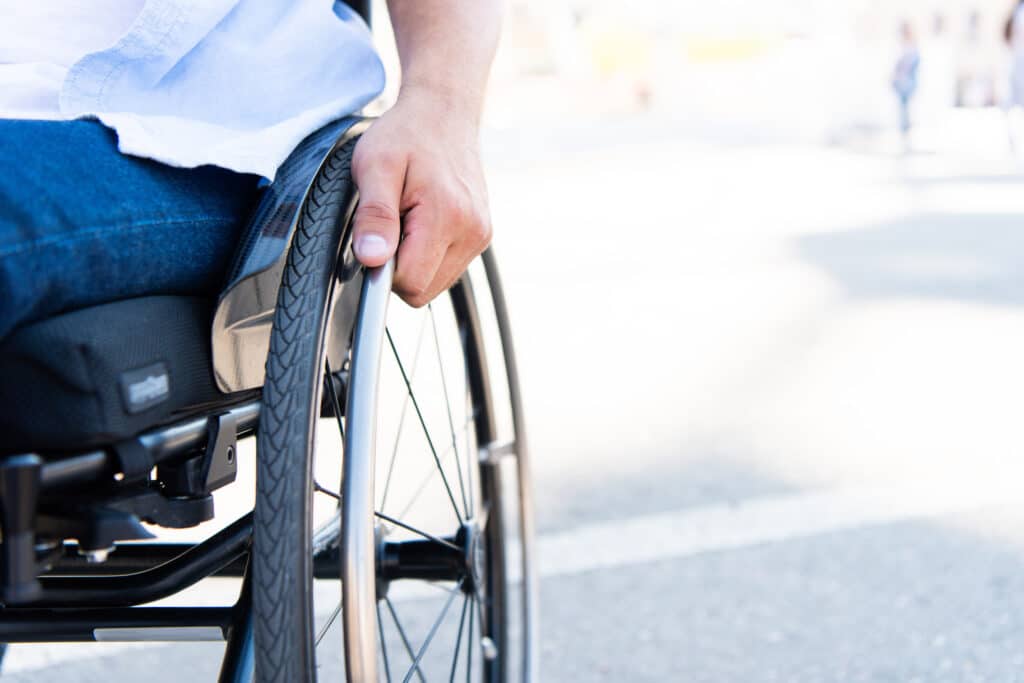 Main d'une personne en fauteuil roulant sur la jante, vêtue d'une chemise bleue et d'un jean, se déplaçant en extérieur.
