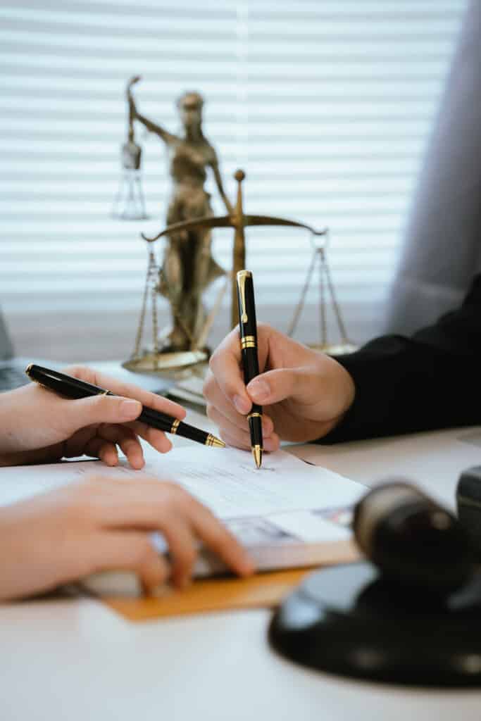 Deux mains signent un contrat avec des stylos noirs devant une statue de la Justice et des balances, symbole légal.