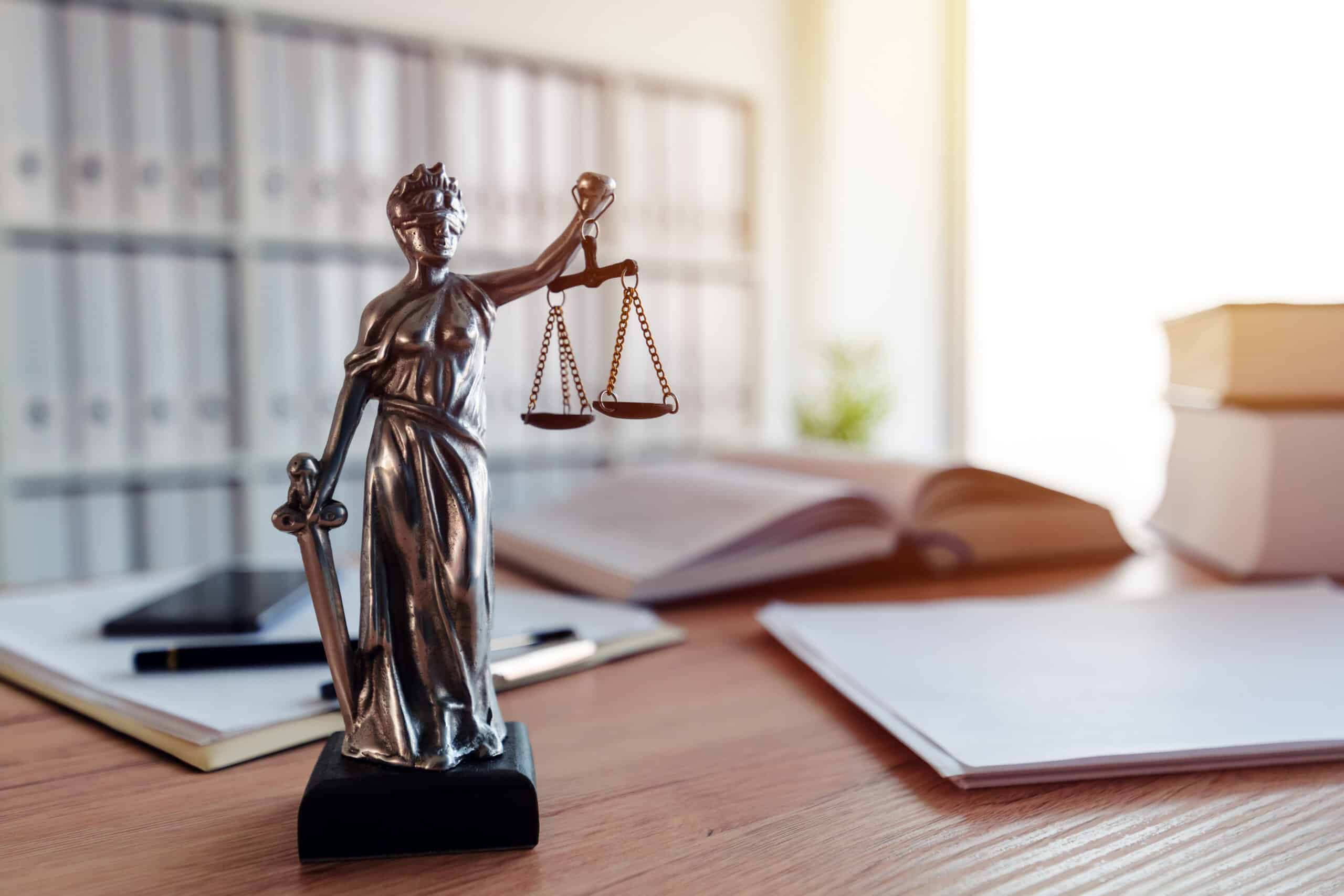 Statue de la Justice blindée tenant épée et balances, posée sur un bureau en bois. En arrière-plan flou, des dossiers et livres de droit.